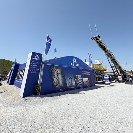 Le stand d'Astec au salon Hillhead 2026 : une structure bleue arborant le logo Astec blanc et gris, située sur un parking en gravier. On aperçoit du matériel Astec derrière le stand.
