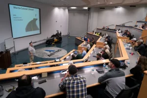 An instructor is teaching a classroom in an auditorium, several rows of students are looking at the instructor and the projectors has a slide that says "What the Customer's Demand" with a road illustration under it.