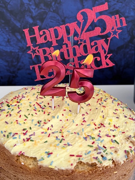 un gâteau d'anniversaire parsemé de paillettes et orné de deux bougies allumées, un « 2 » et un « 5 », ainsi qu'une pancarte rouge découpée sur laquelle on peut lire « Joyeux 25e anniversaire, Telestack »